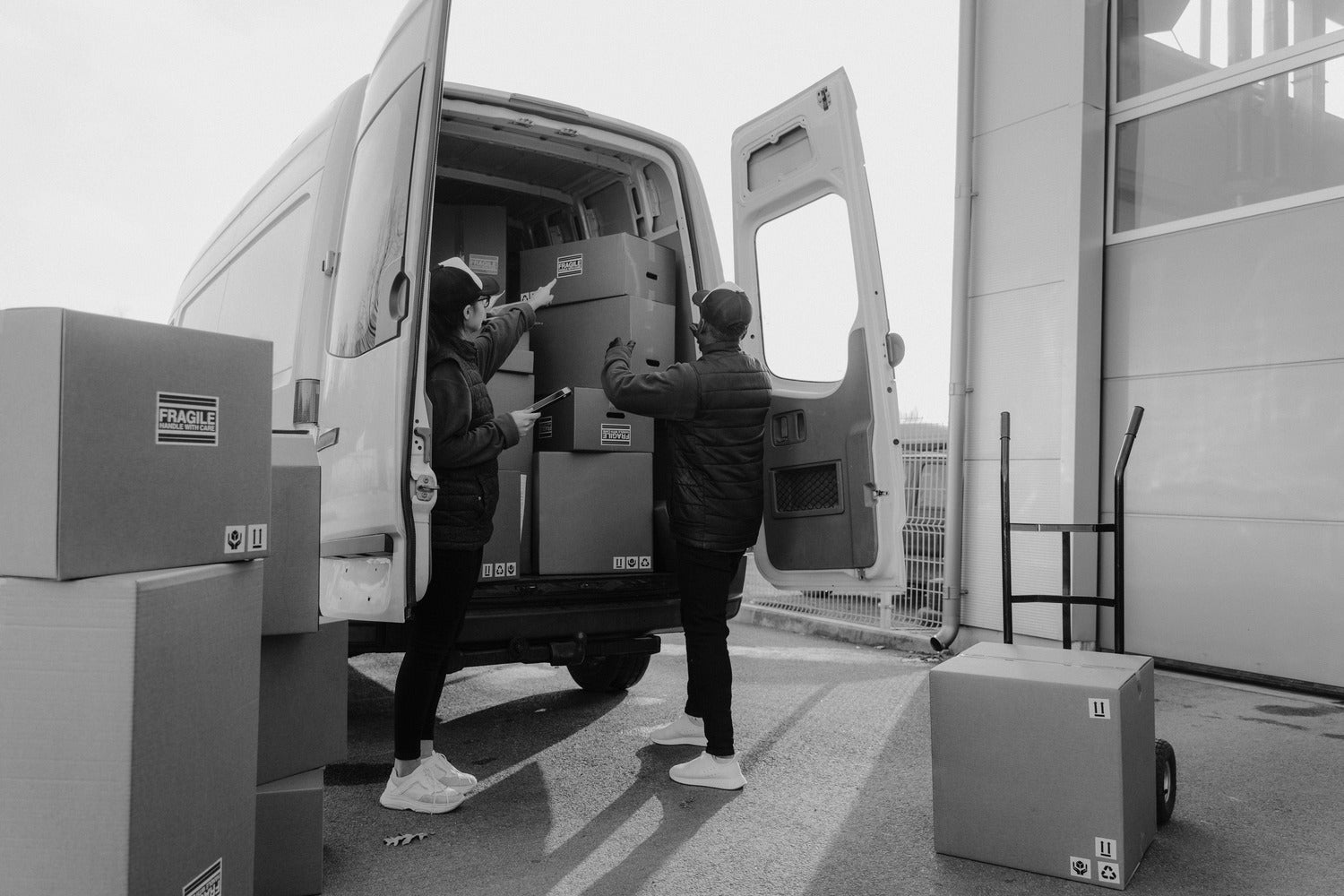 Two people loading large boxes into a van with a warehouse in the background.